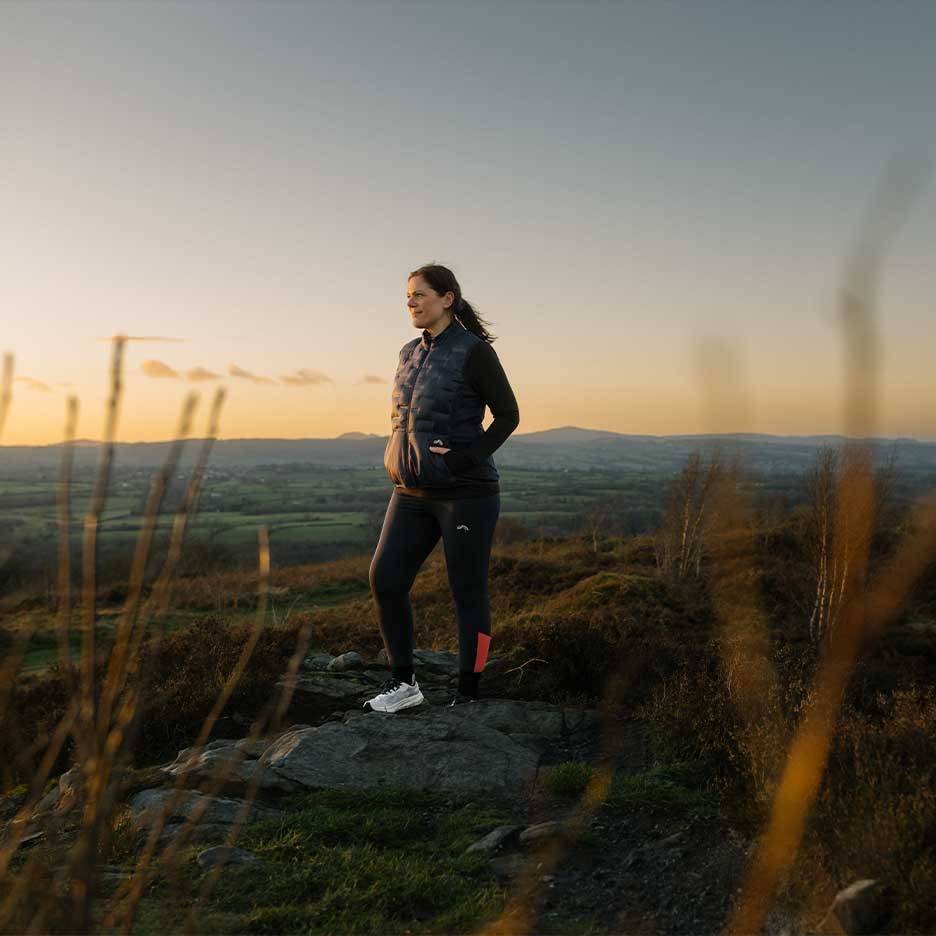 Donna con gilet trapuntato e leggings in piedi su una collina al tramonto.