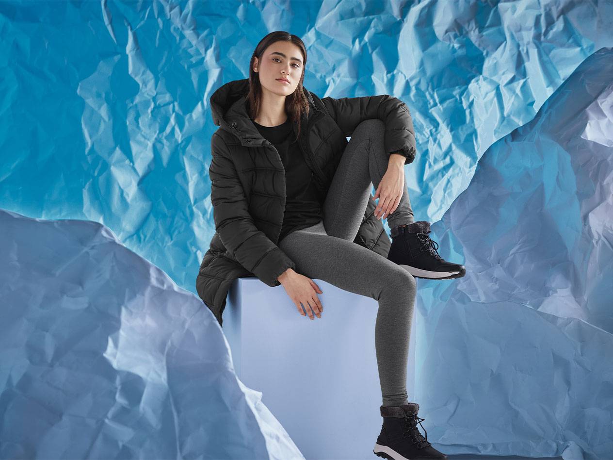 Donna in piumino nero, leggings grigi e stivali invernali neri seduta su una scatola blu.
