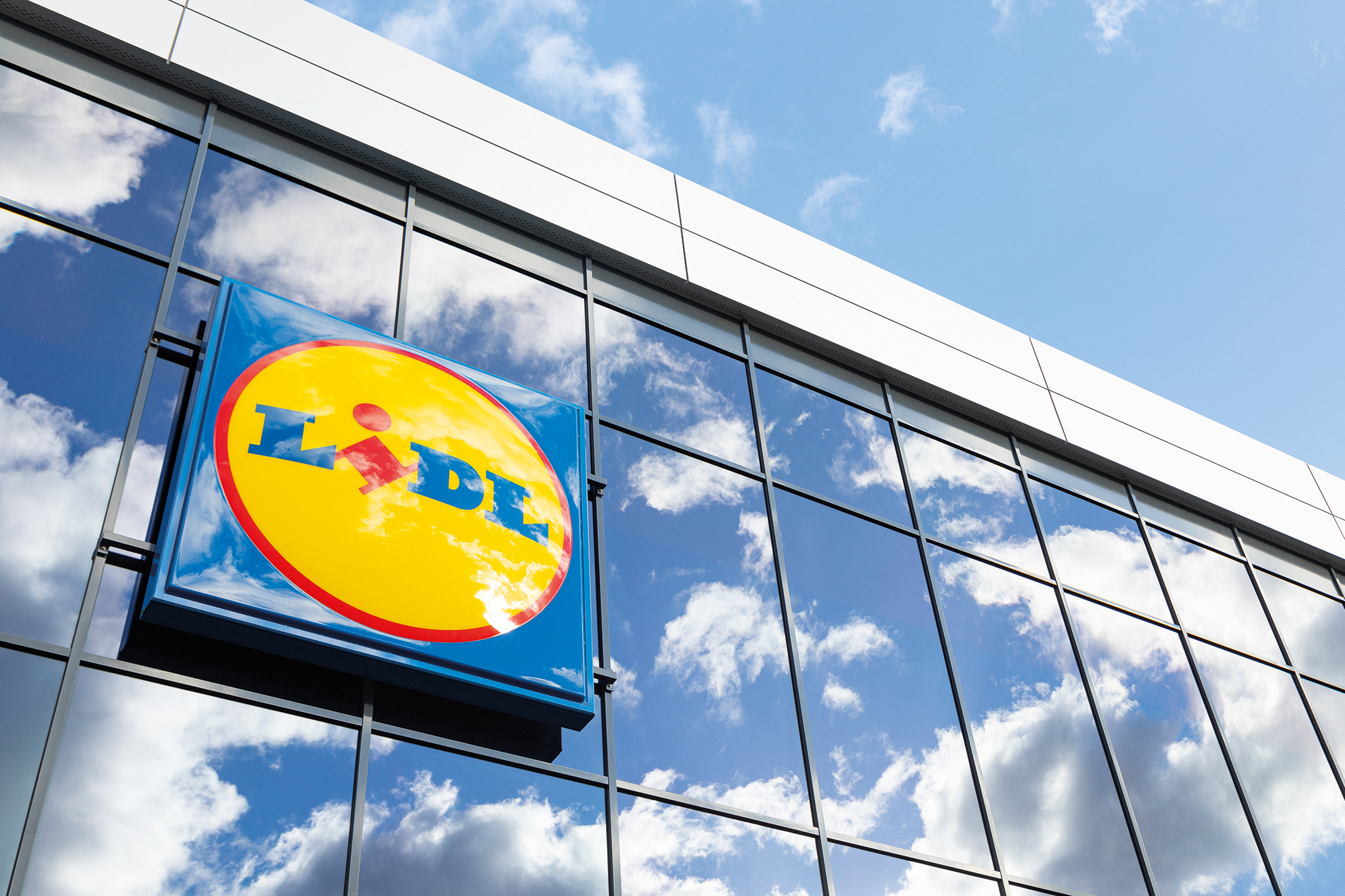 Insegna Lidl su facciata a vetri di un edificio, con riflesso di cielo e nuvole.