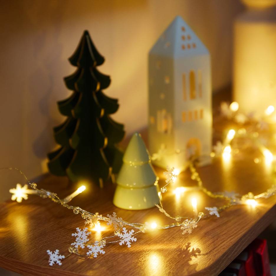 Decorazioni natalizie: un albero verde scuro, un albero verde chiaro, una casa bianca e una catena luminosa con fiocchi di neve.