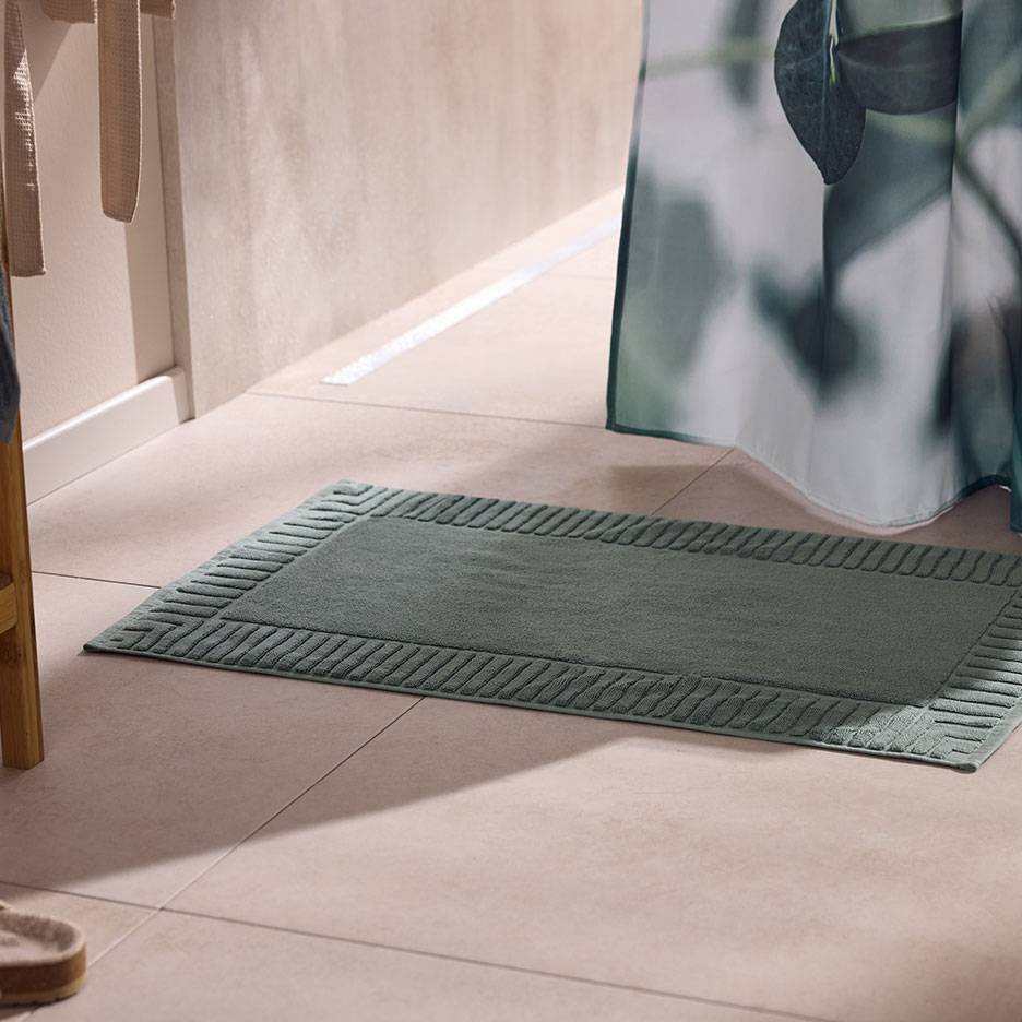 Tappetino da bagno verde testurizzato su piastrelle chiare, accanto a una tenda da doccia.