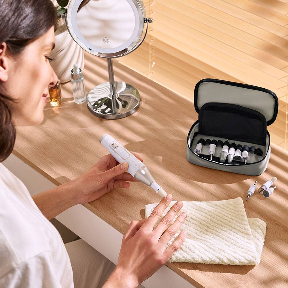 Donna usa lima unghie elettrica con accessori, accanto a specchio e custodia con strumenti.