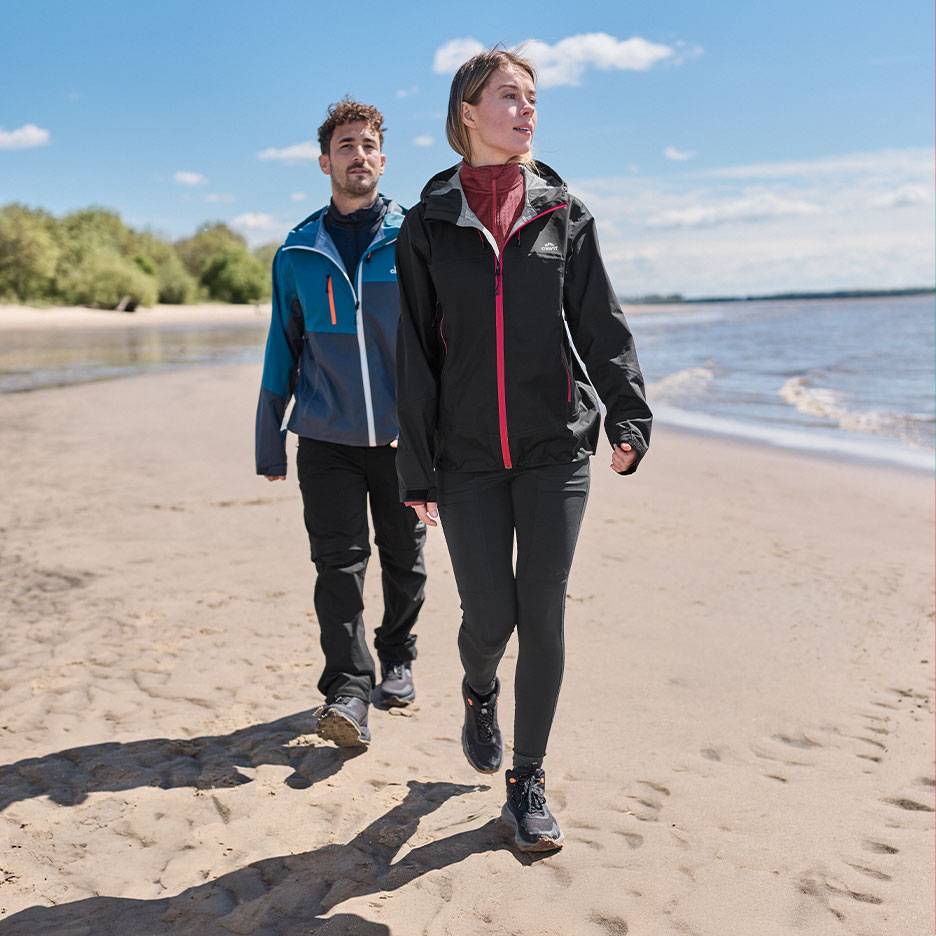 Coppia in giacche e pantaloni da trekking cammina su una spiaggia sabbiosa in riva al mare.