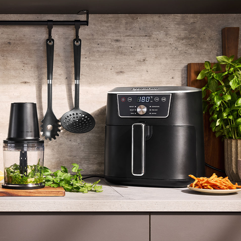 Friggitrice ad aria SilverCrest e tritatutto su un bancone della cucina con erbe fresche e patatine fritte di patate dolci.