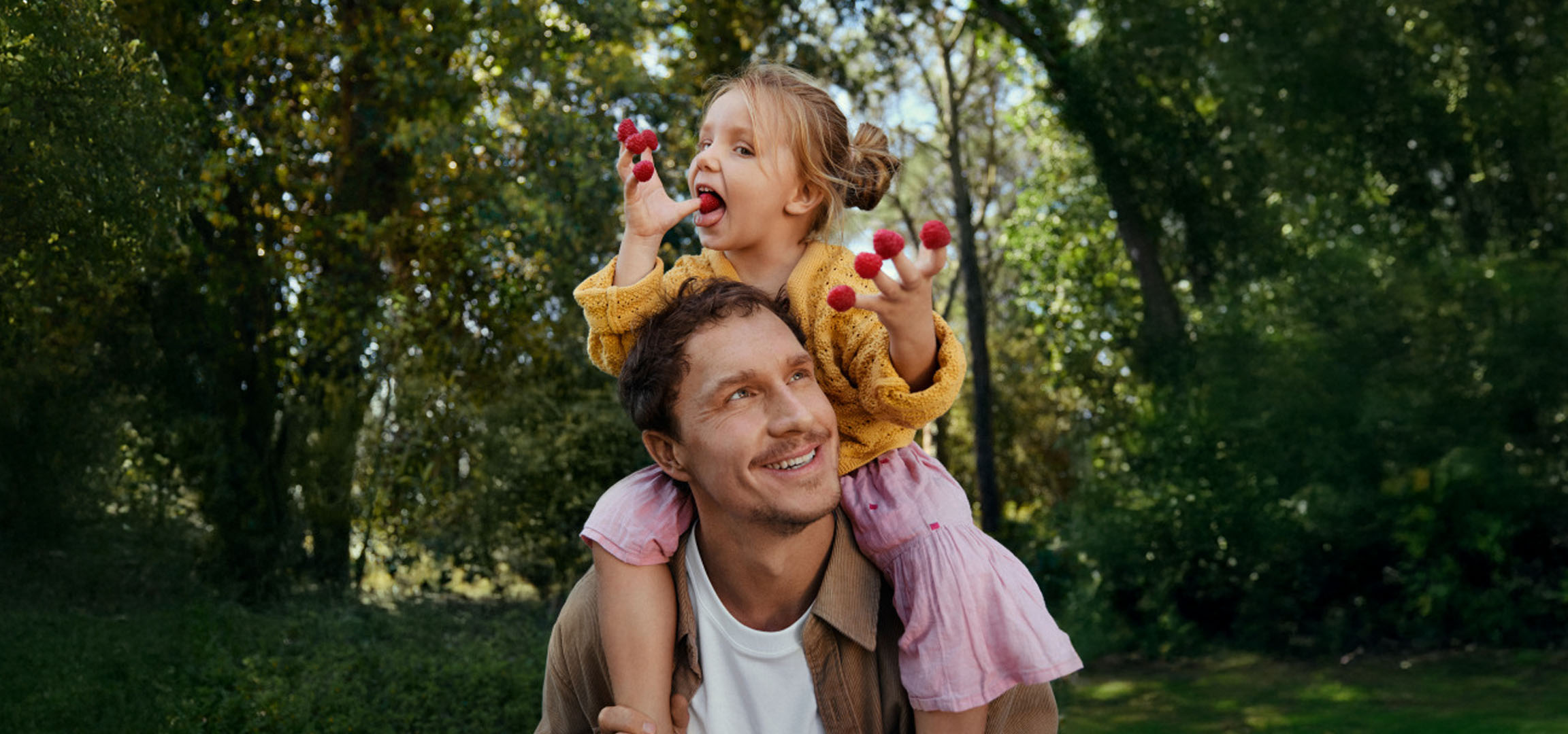 Un padre sorridente porta una bambina sulle spalle, che mangia lamponi in un bosco.