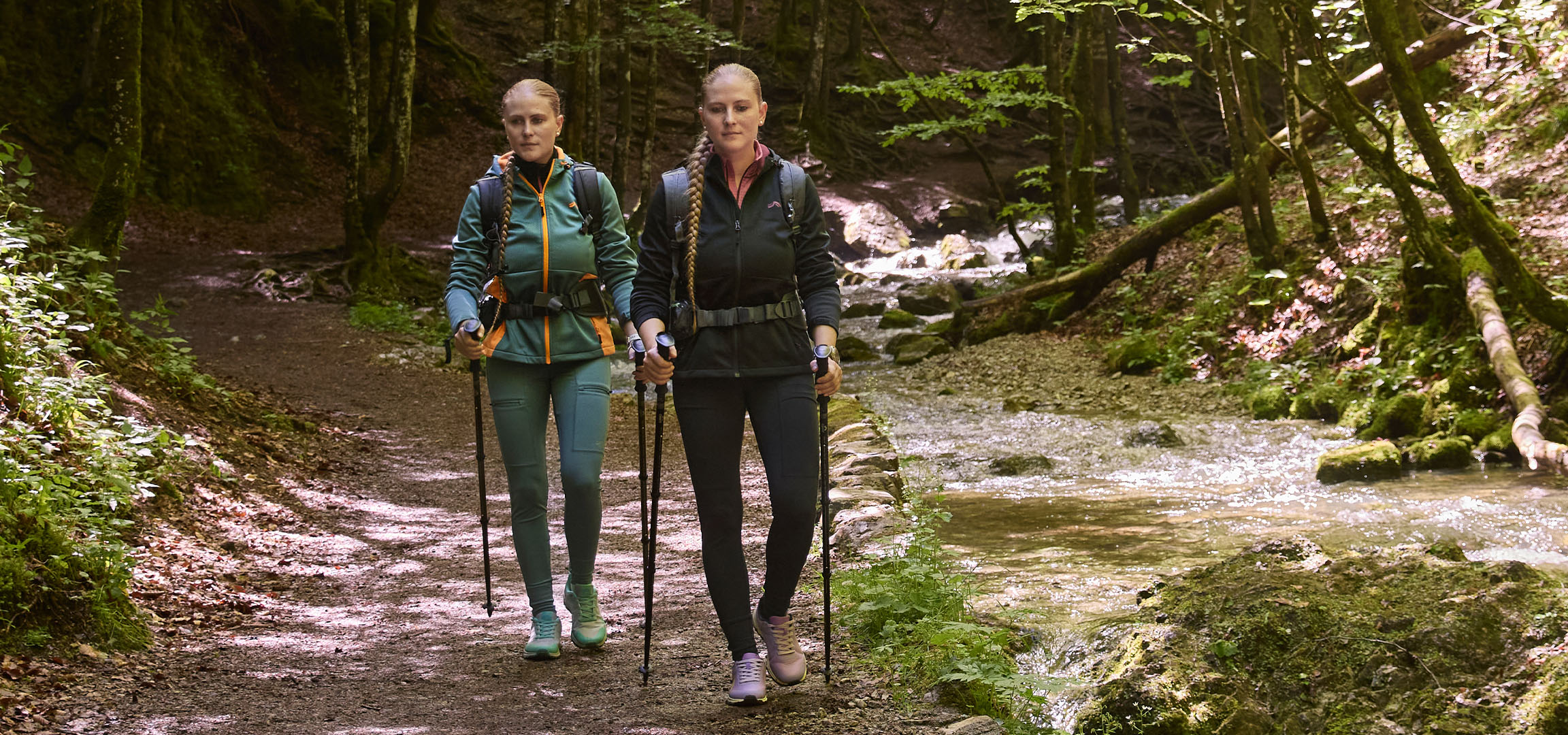 Due donne camminano lungo un ruscello in un bosco con i bastoncini da trekking.