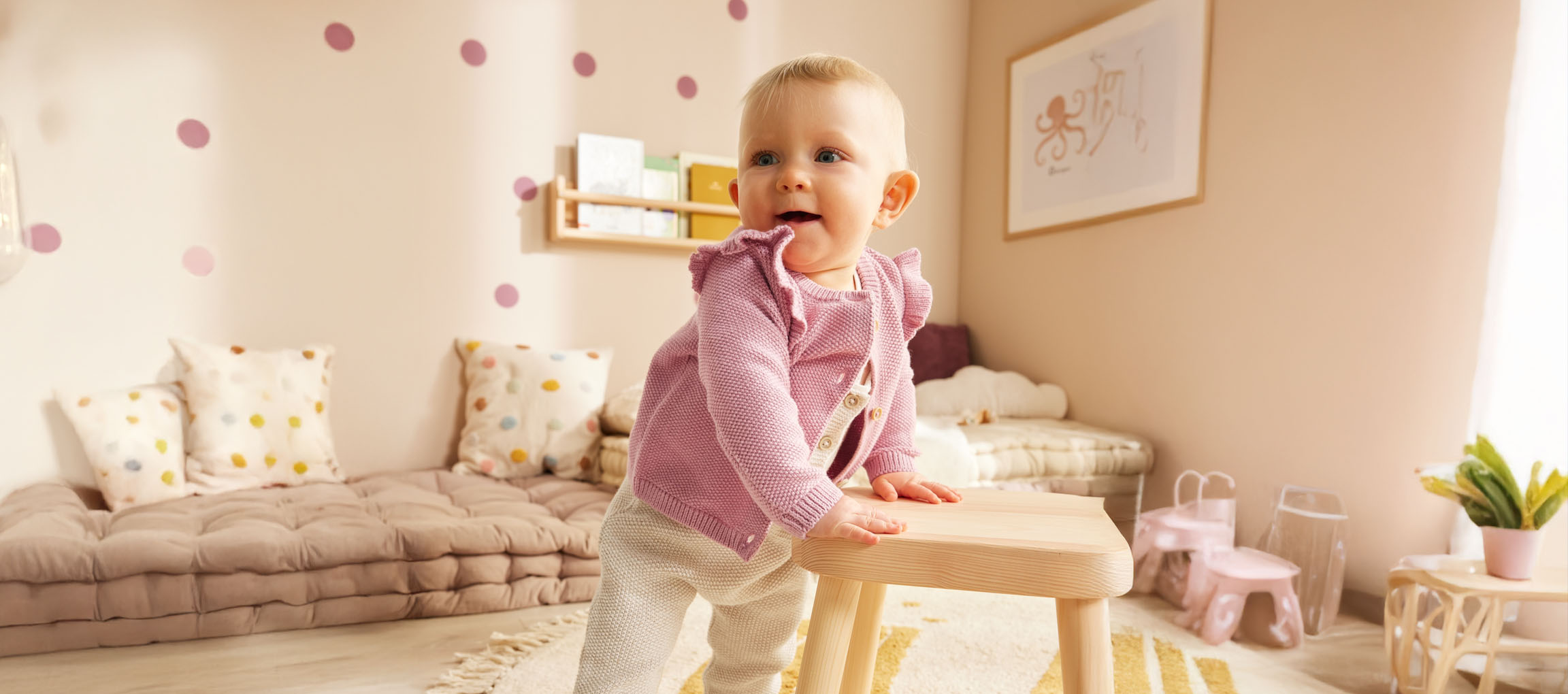 Una bambina in un carino cardigan rosa si alza in piedi vicino a uno sgabello di legno nella sua cameretta.