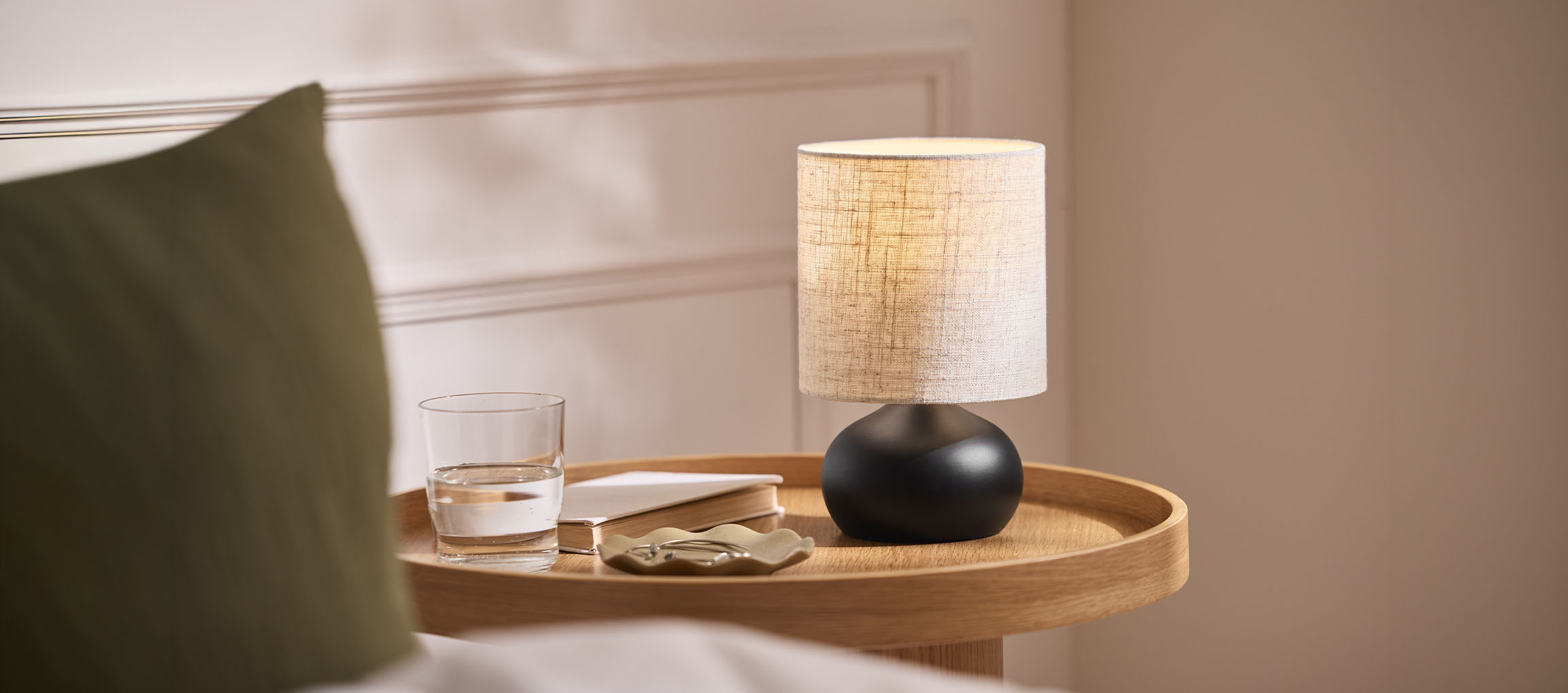 Lampada da comodino con paralume in tessuto, bicchiere d'acqua e libro su vassoio di legno.