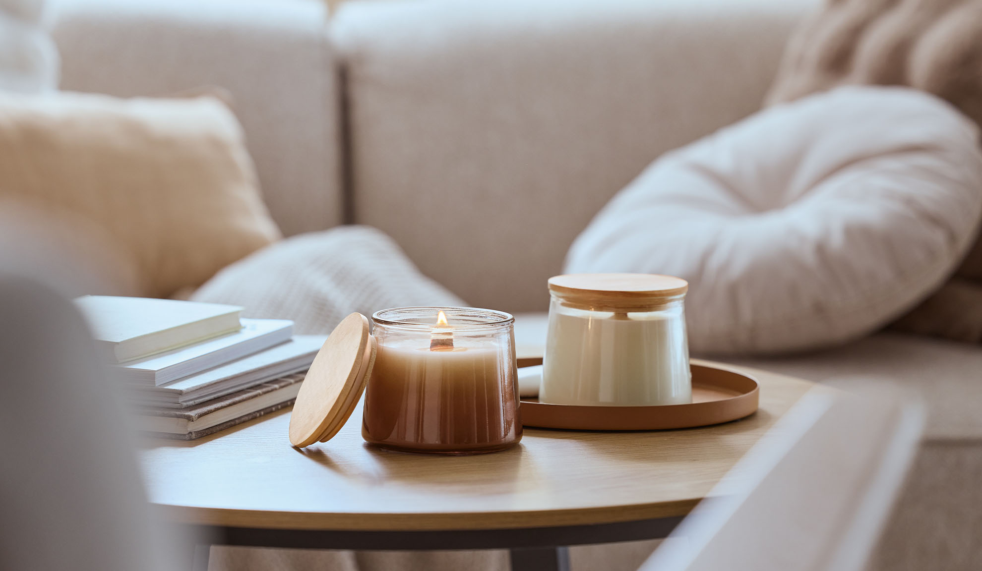 Due candele profumate su un tavolo di legno, una accesa, con un divano accogliente sullo sfondo.