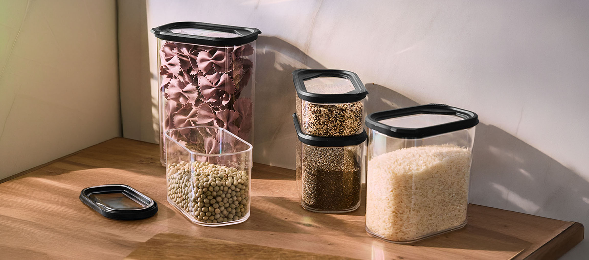 Contenitori da cucina trasparenti con coperchi neri, pieni di pasta, lenticchie, quinoa, semi di chia e riso su una superficie di legno.