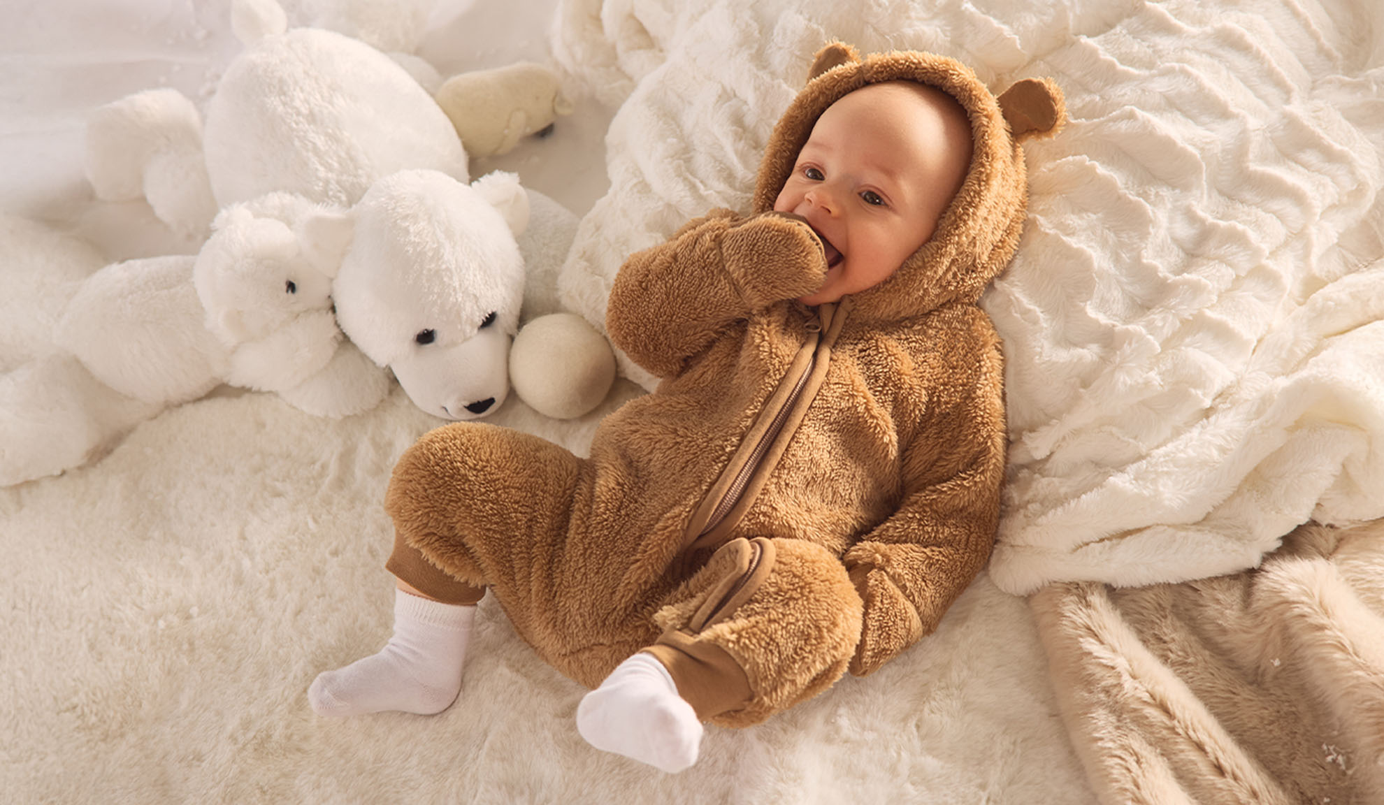 Bambino in tutina da orso marrone, sdraiato su una coperta bianca soffice con orsetti polari giocattolo.