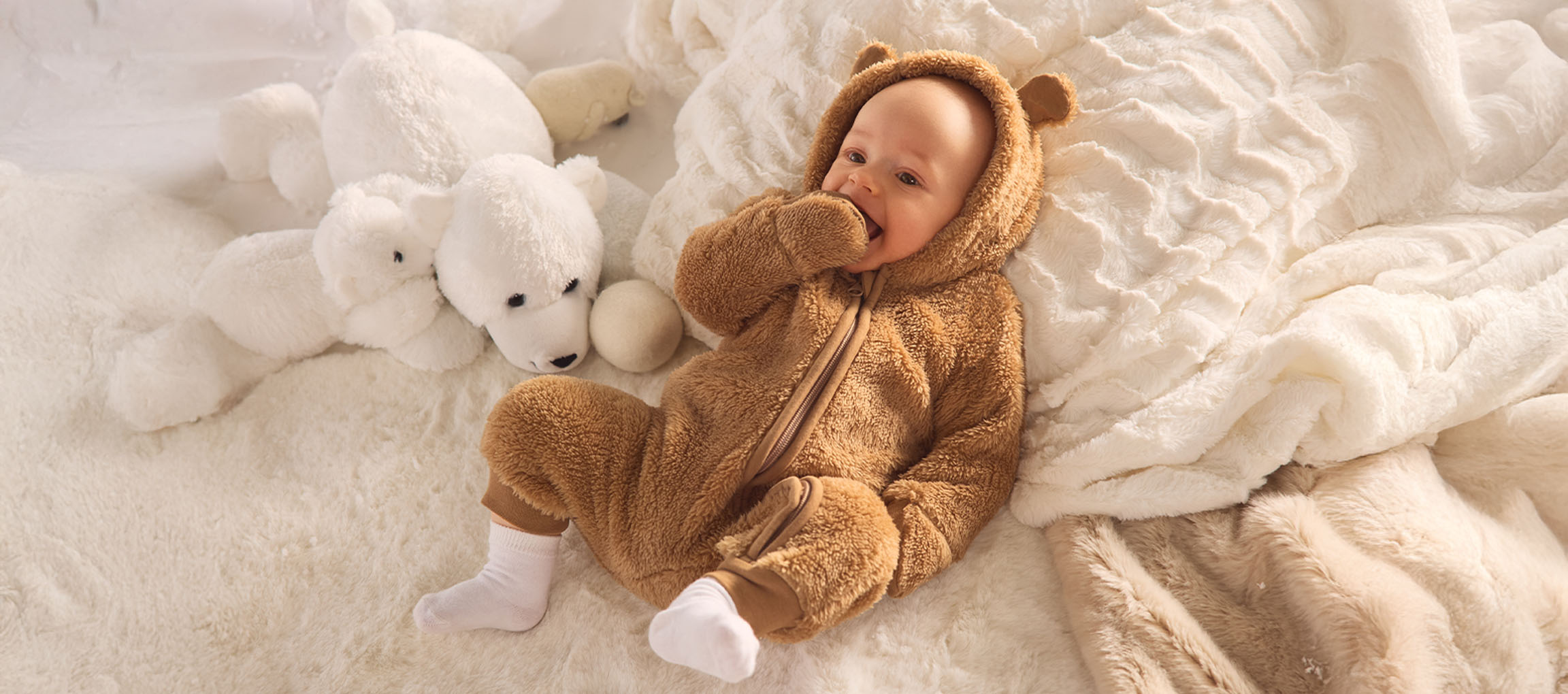 Bambino in tutina da orso marrone, sdraiato su una coperta bianca e soffice con orsetti polari.