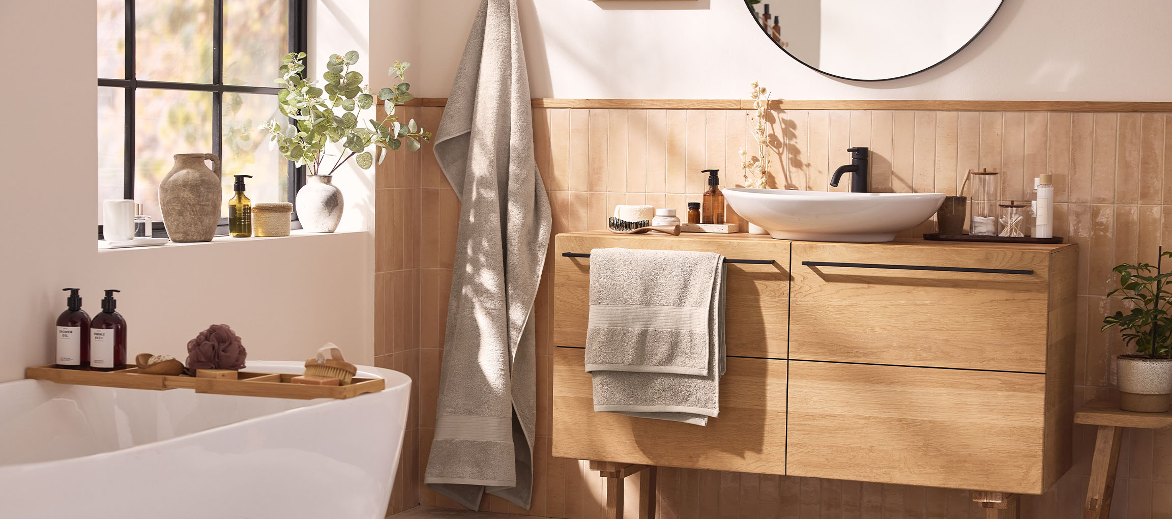 Un bagno moderno con mobile in legno, lavabo bianco, specchio rotondo e vasca da bagno con accessori.