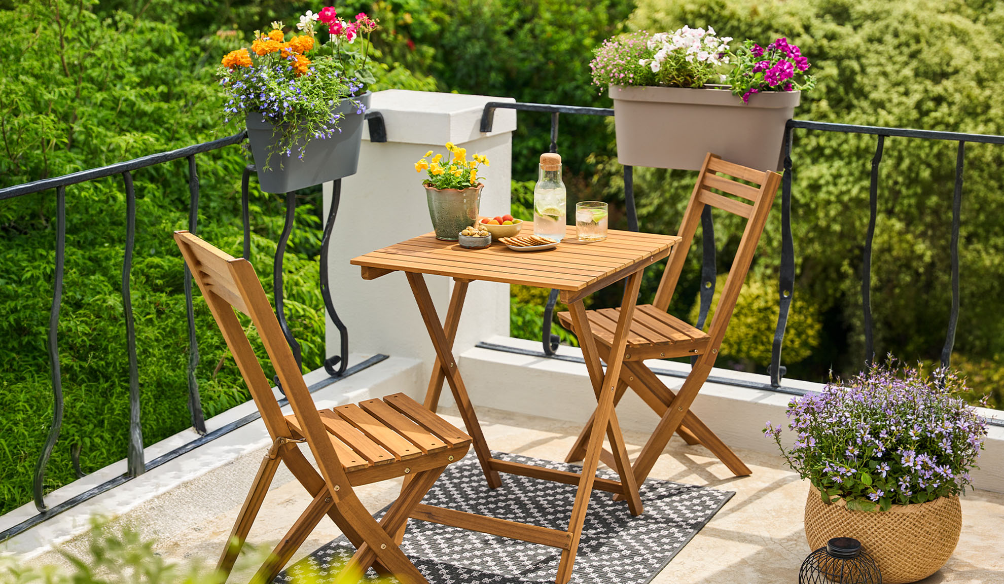 Balcone con set bistrò in legno, fiori in vaso e bevande su un tappeto a fantasia.