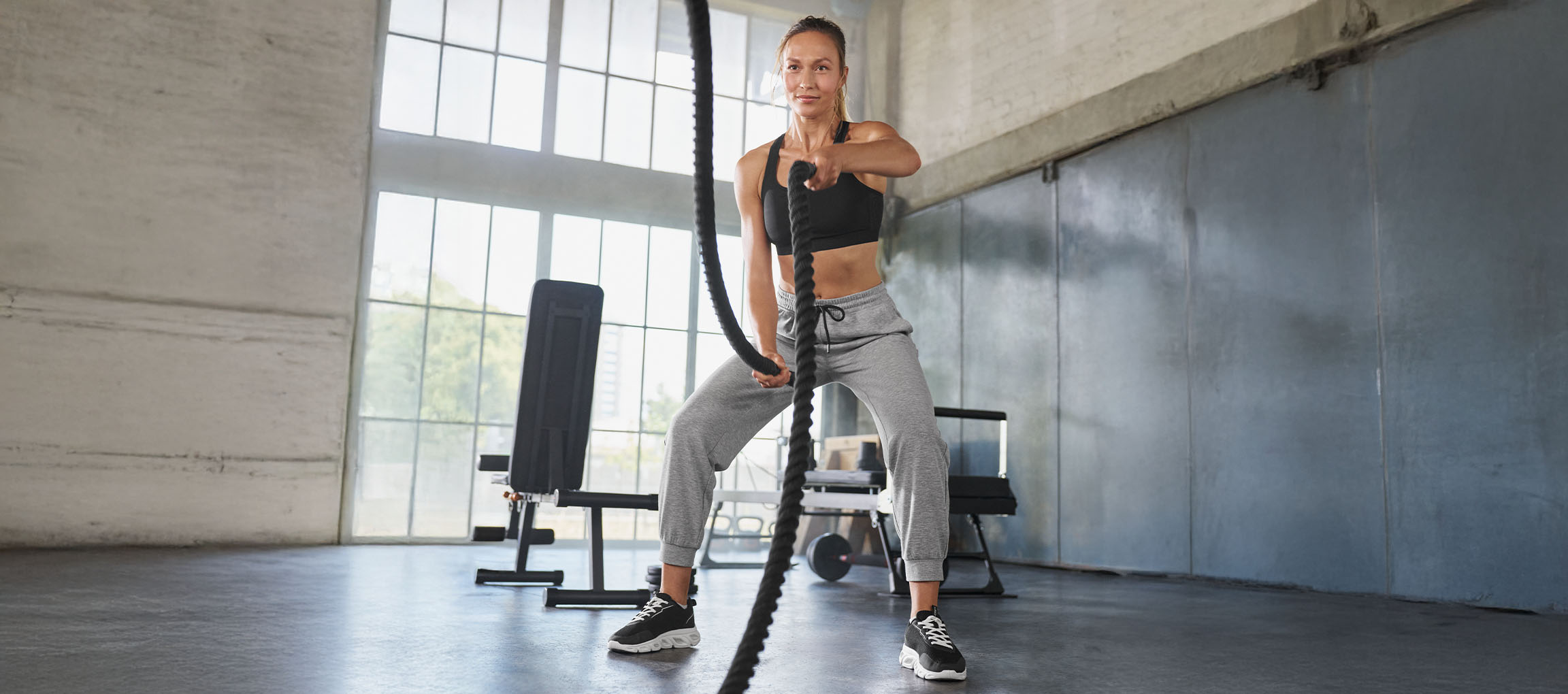 Donna in reggiseno sportivo nero e pantaloni della tuta grigi si allena con le battle ropes
