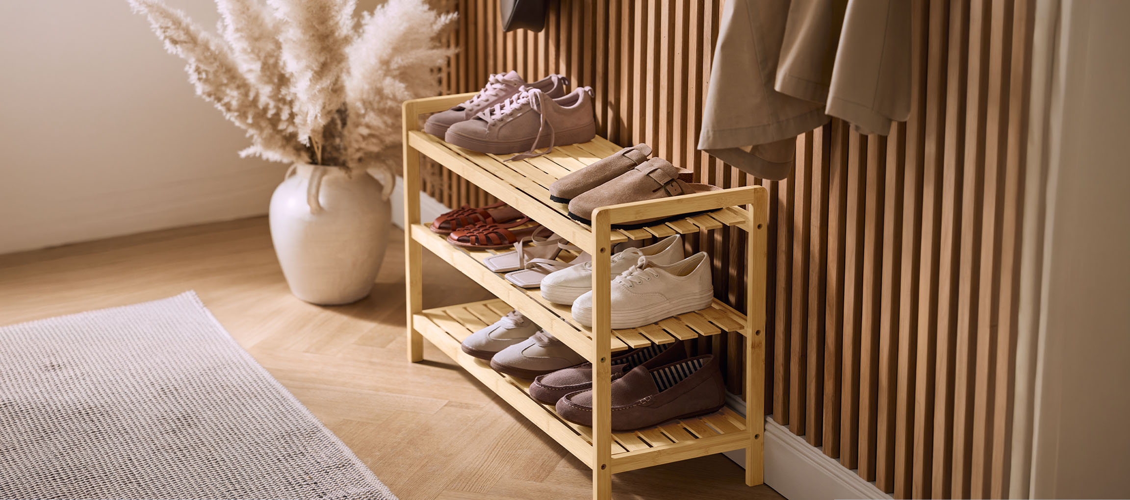 Scarpiera in legno con varie scarpe, un vaso di erba della pampa e un cappotto sulla parete.
