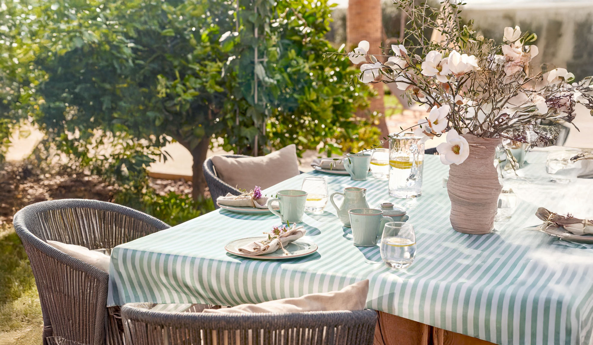 Tavolo da pranzo all'aperto con tovaglia a righe, stoviglie in ceramica e vaso di fiori.