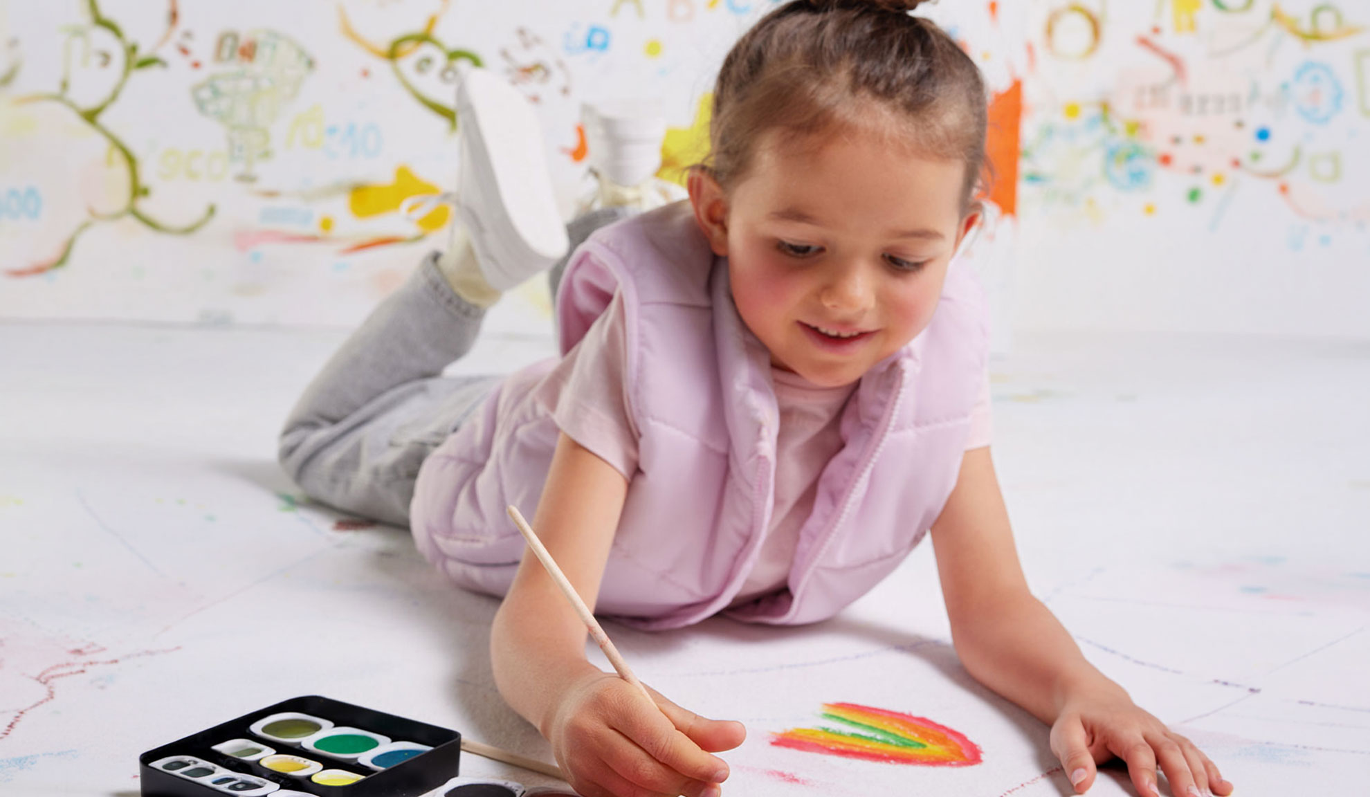 Una bambina dipinge un arcobaleno su carta con acquerelli, indossando un gilet rosa.