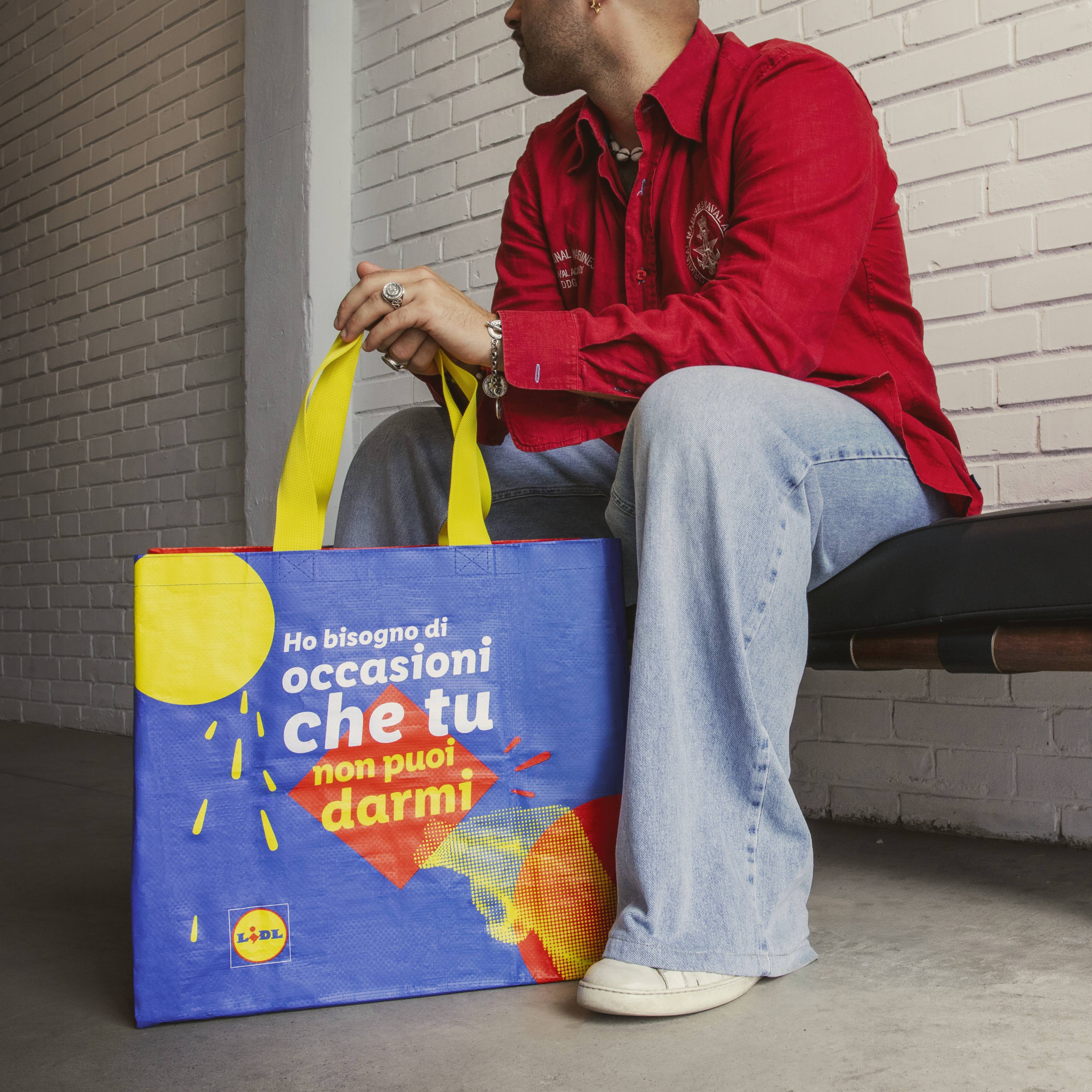 Un uomo siede vicino a una borsa della spesa Lidl con la scritta 'Ho bisogno di occasioni che tu non puoi darmi'.