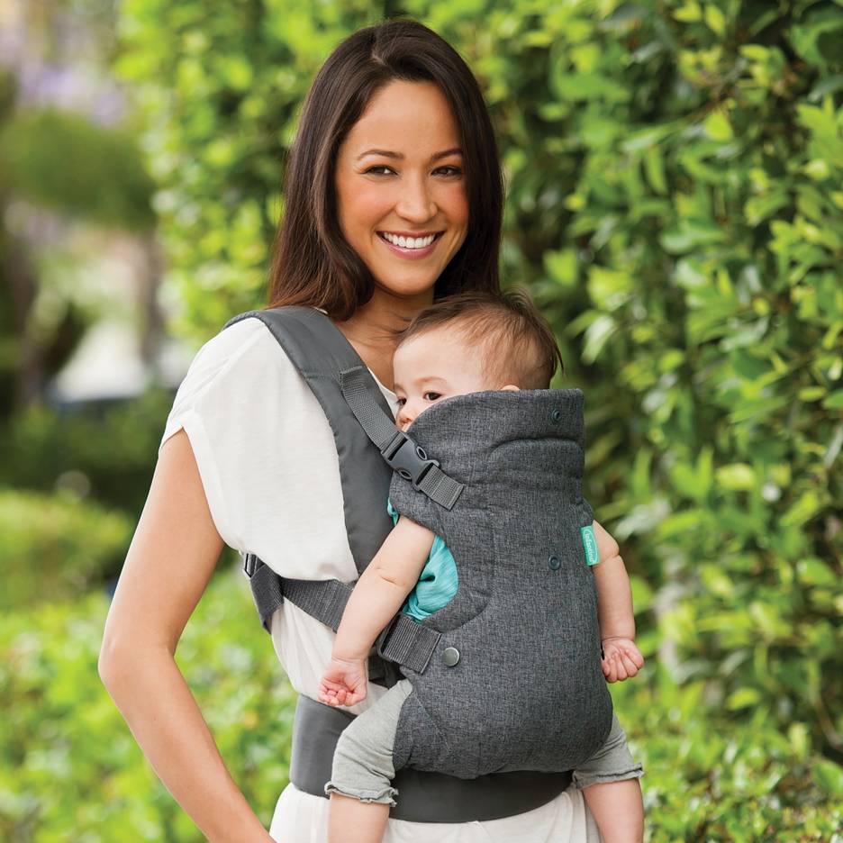 Donna sorridente con marsupio grigio che porta un bambino all'aperto.