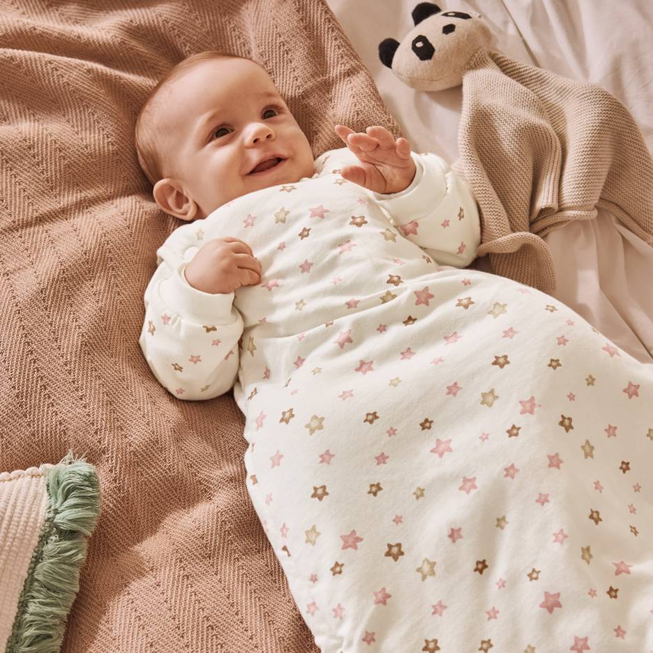 Bebè sorridente in sacco nanna bianco con stelle rosa e marroni, sdraiato su una coperta.