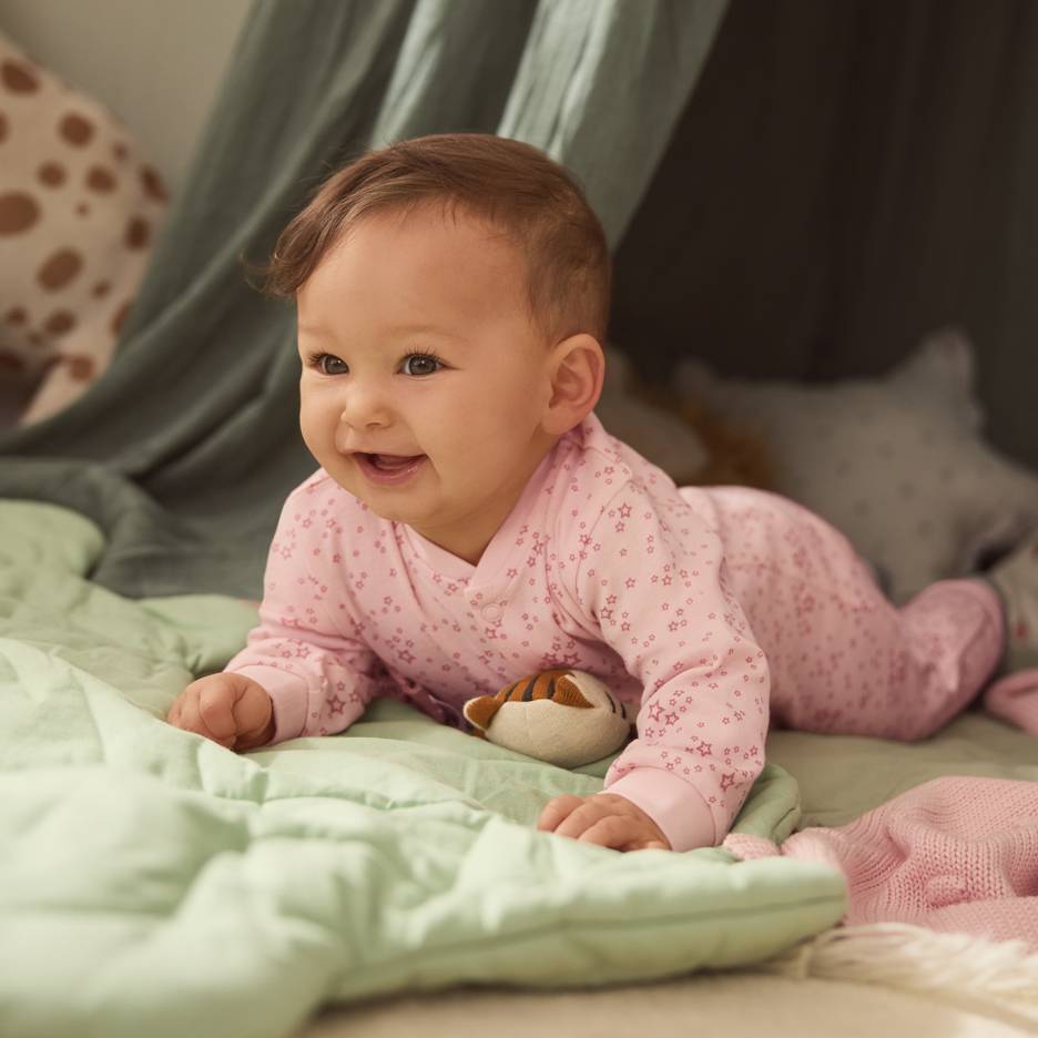 Bebè in pigiama rosa a stelle, sdraiato su una coperta verde con un peluche.