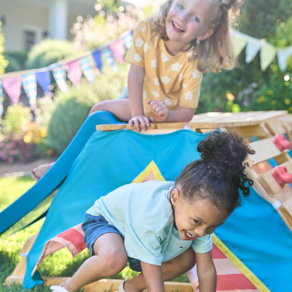 Due bambini felici giocano su una struttura da arrampicata in legno con una tenda blu in un giardino soleggiato.