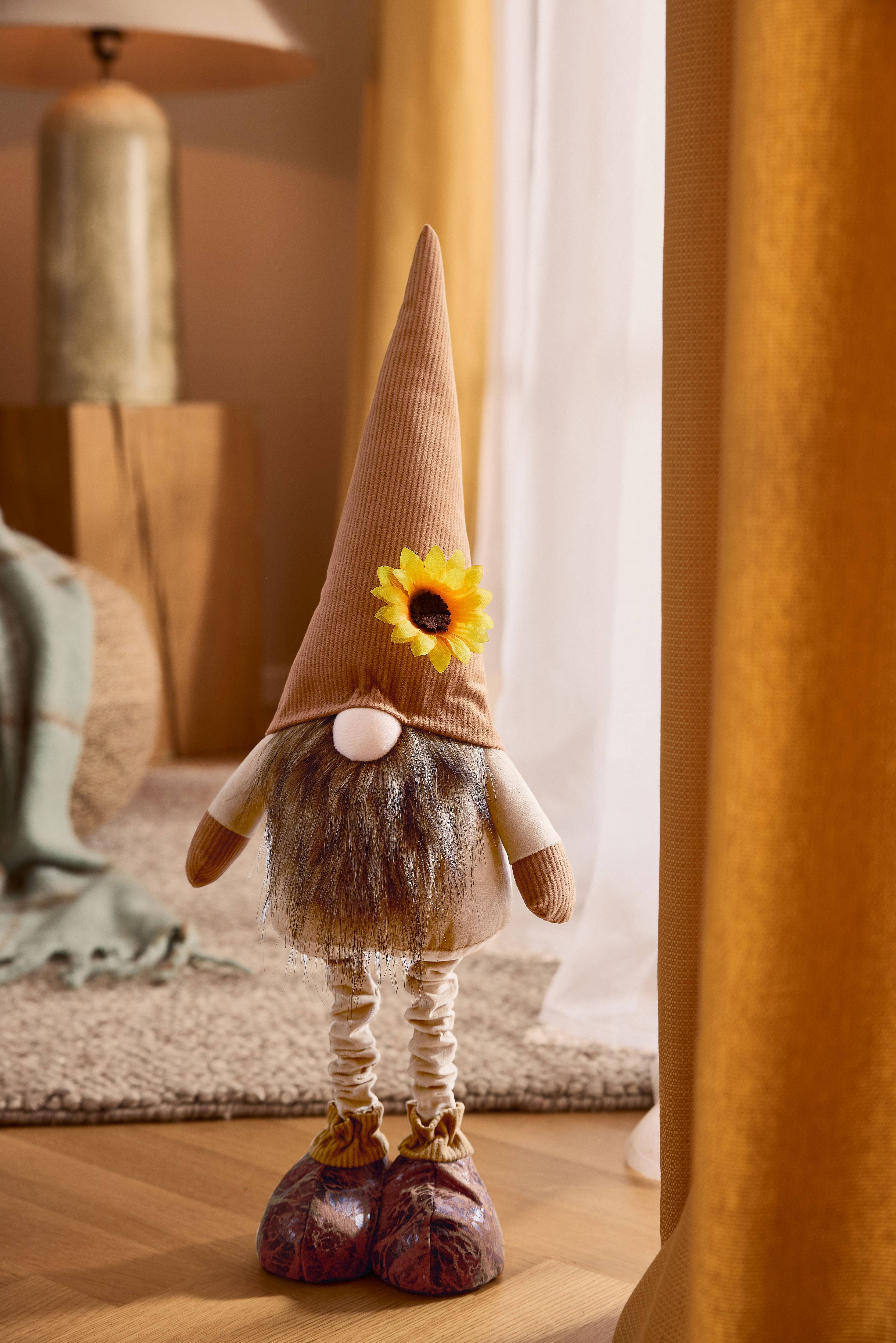 Gnomo di peluche con cappello a punta marrone, barba e girasole, in un ambiente accogliente.