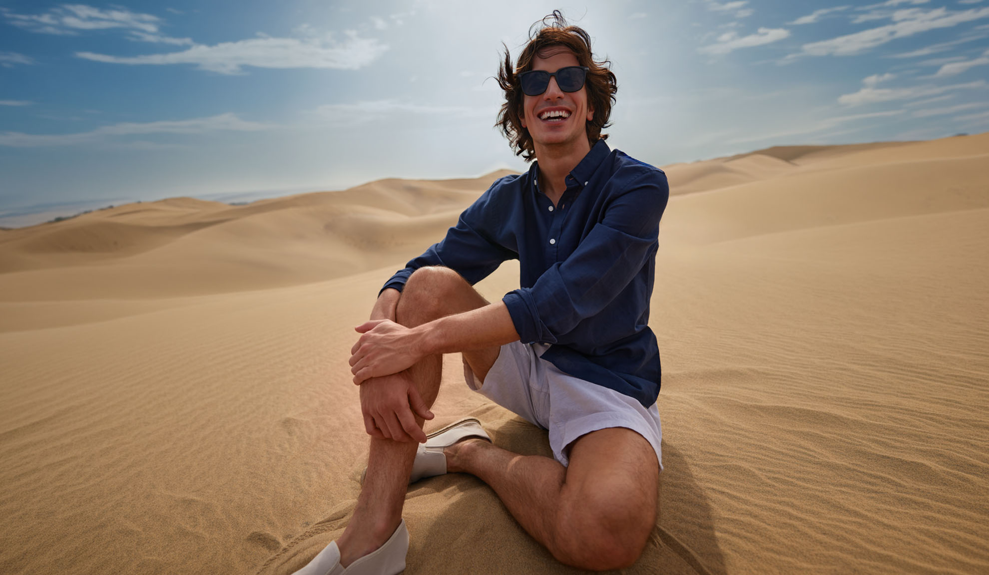 Uomo in camicia blu scuro e pantaloncini chiari seduto sulle dune di sabbia, sorridendo.