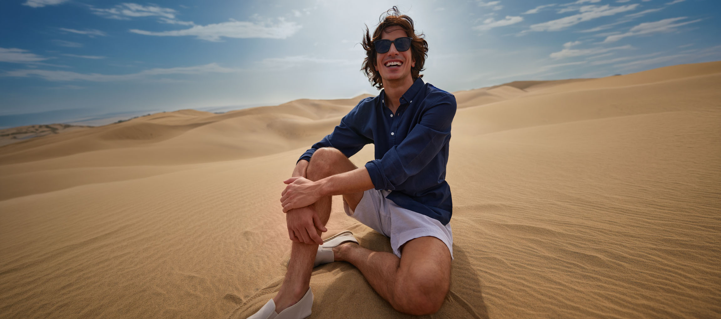 Uomo in camicia blu scuro e pantaloncini seduto sulle dune di sabbia, sorridente.