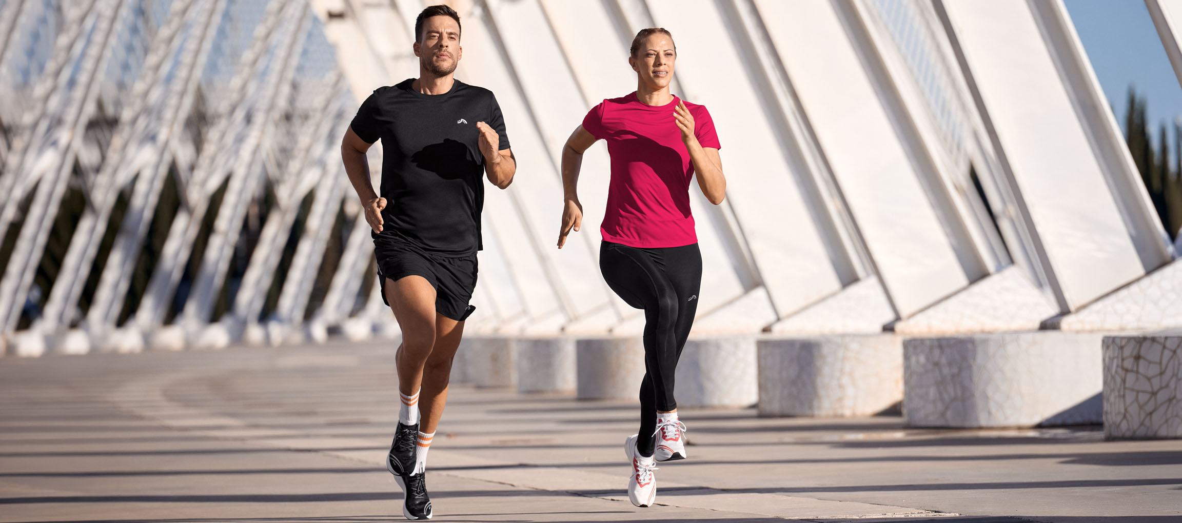 Uomo e donna corrono in abbigliamento sportivo, top neri e rosa, pantaloncini e leggings neri.