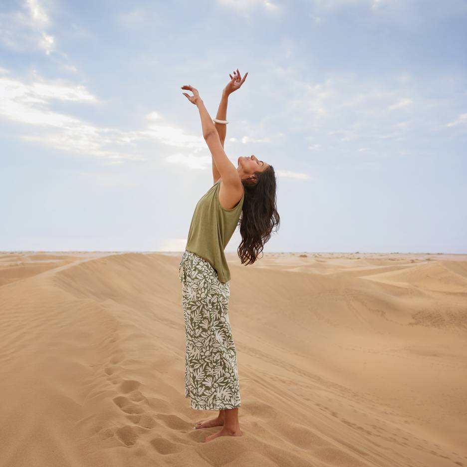 Donna tra le dune di sabbia con top verde e pantaloni fantasia, braccia alzate.