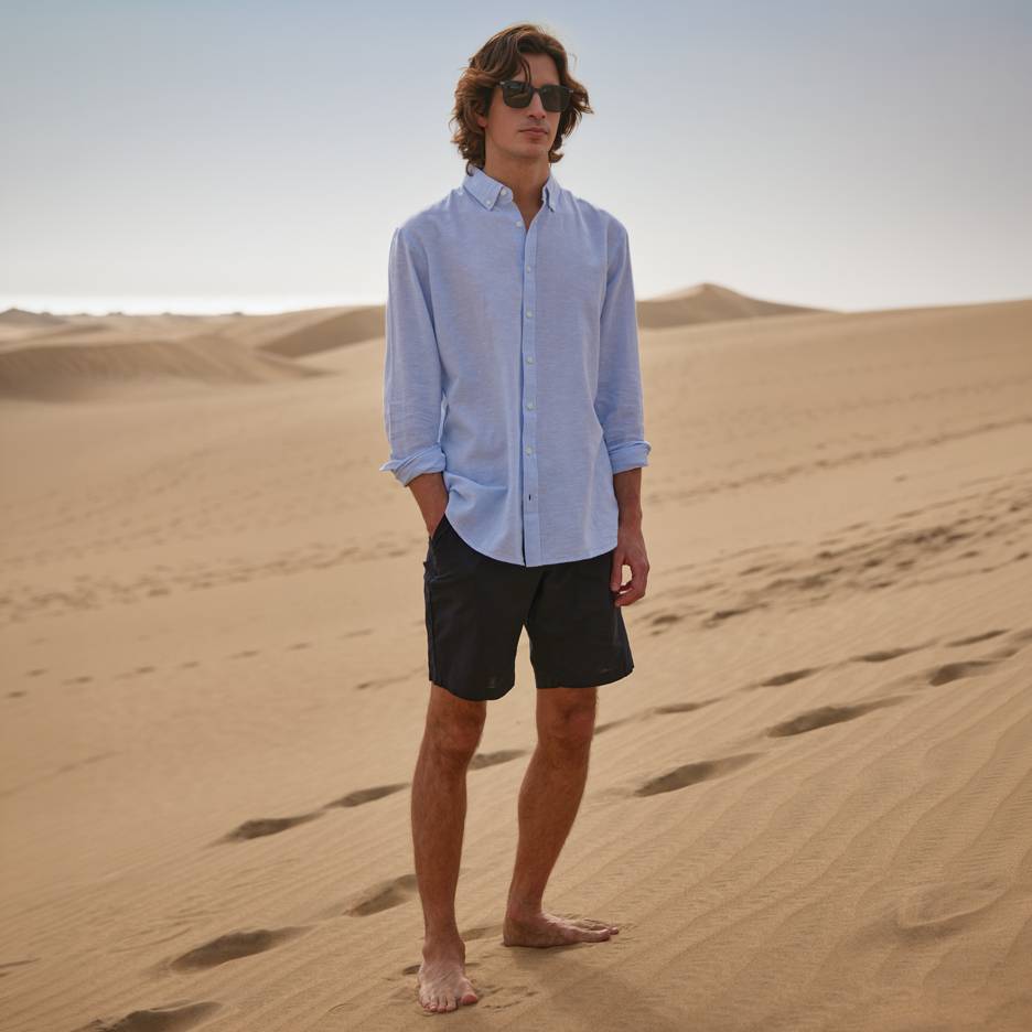 Uomo in camicia blu e pantaloncini neri, a piedi nudi sulle dune di sabbia.