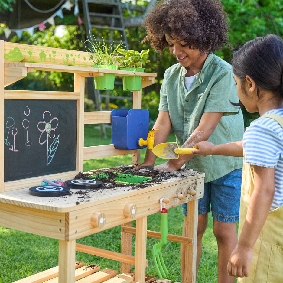Due bambini giocano con una cucina di fango in legno, con lavagna e piante in vaso.