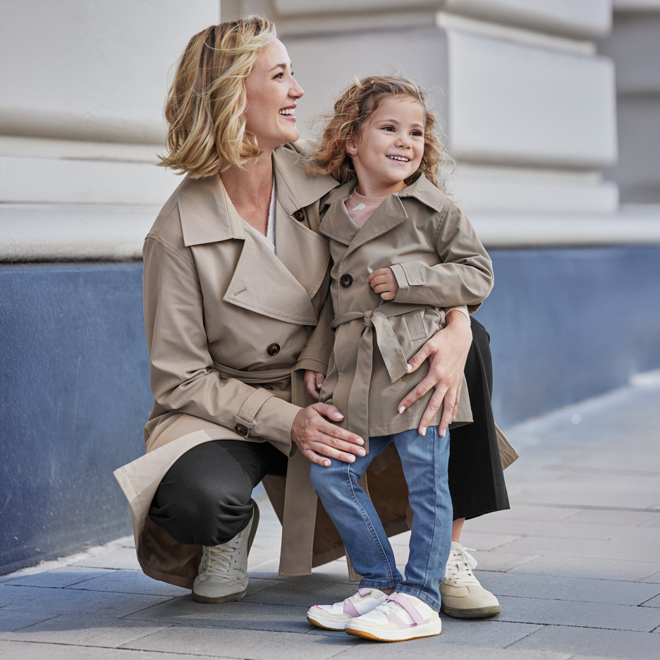 Due donne in trench beige, un'adulta e una bambina, sorridono all'aperto.
