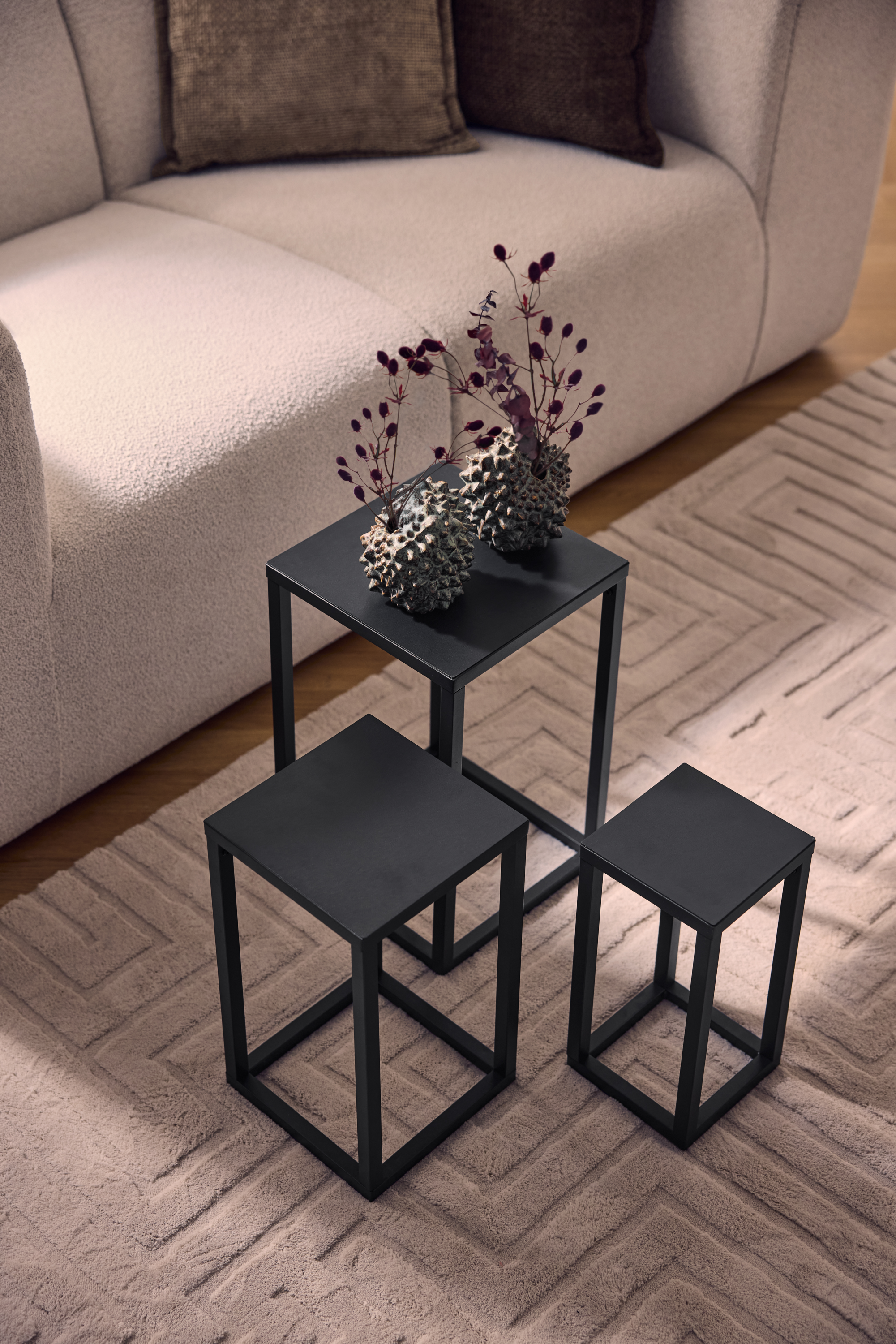 Tre tavolini da caffè in metallo nero su un tappeto beige con motivo geometrico, accanto a un divano chiaro.