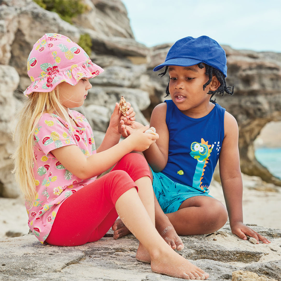 Due bambini in spiaggia, uno con cappello da sole rosa e top a fantasia di frutta, l'altro con cappellino blu e canotta con dinosauro, che tengono conchiglie.