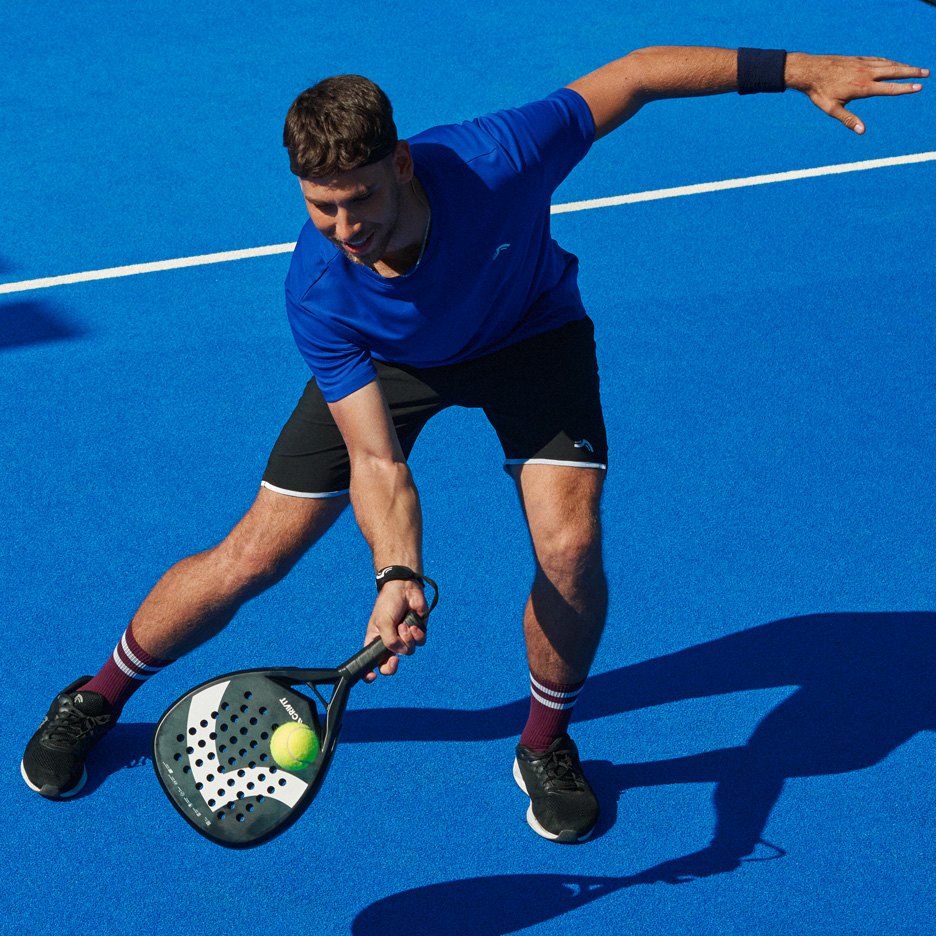 Uomo in maglietta blu e pantaloncini neri gioca a padel con racchetta e palla.
