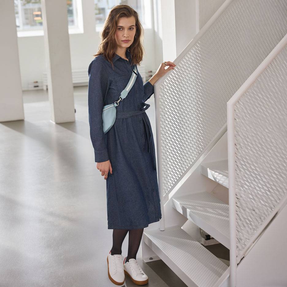 Donna in vestito camicia di jeans, calze nere e sneakers bianche, con marsupio azzurro.