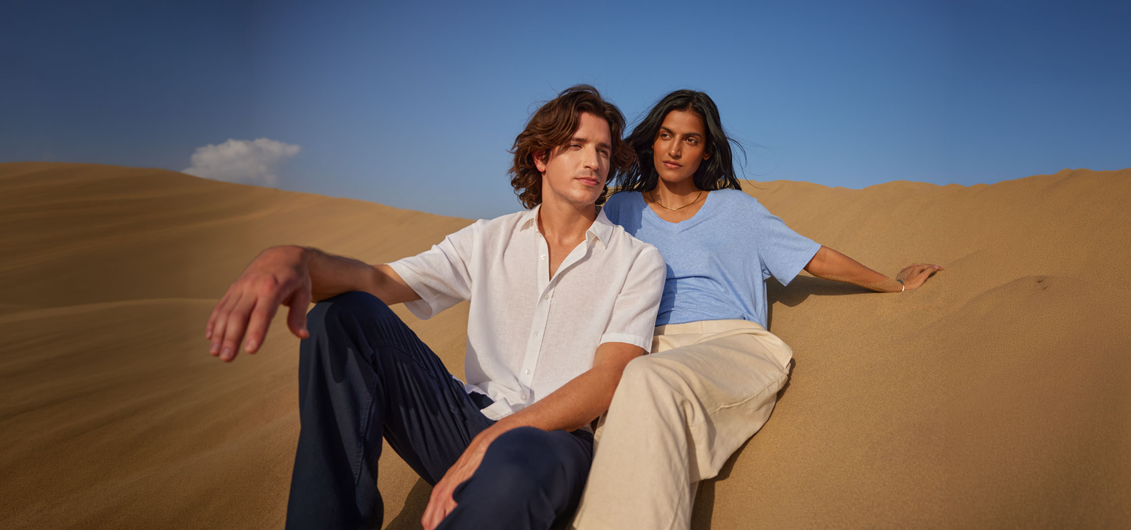 Uomo e donna seduti sulle dune di sabbia, indossando camicie e pantaloni di lino.