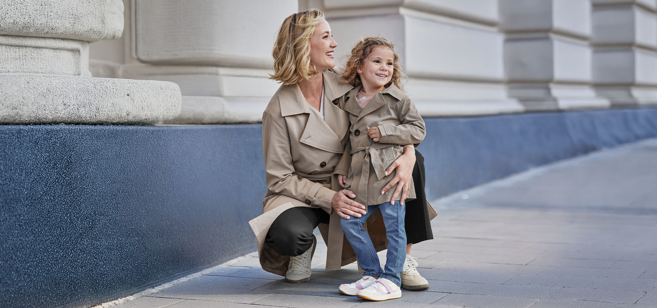 Due donne in trench beige, un'adulta e una bambina, sorridono all'aperto.