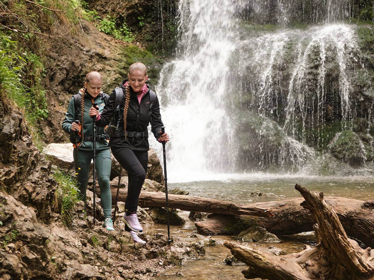 Due donne con bastoni da trekking camminano su un sentiero roccioso vicino a una cascata.