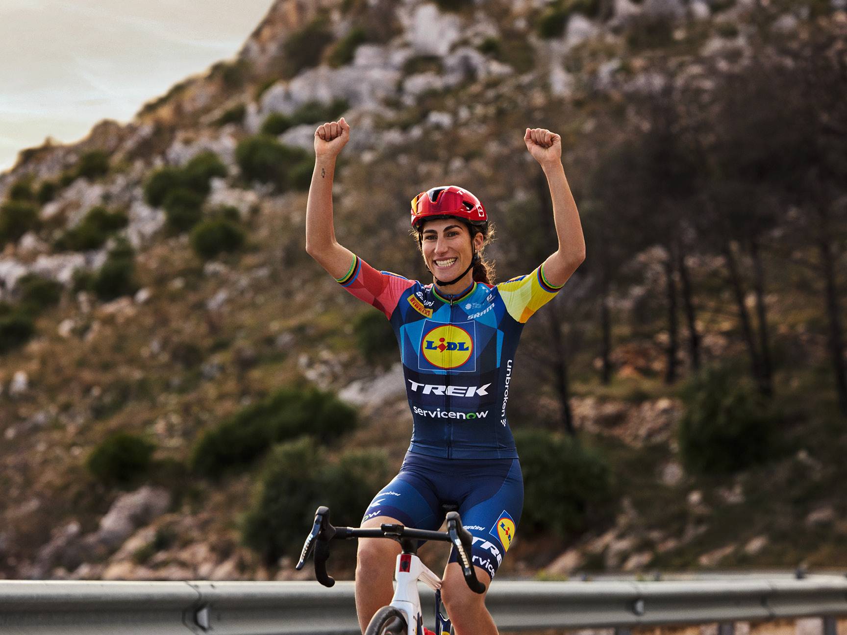 Ciclista donna sorridente con maglia e pantaloncini blu, braccia alzate in segno di vittoria.