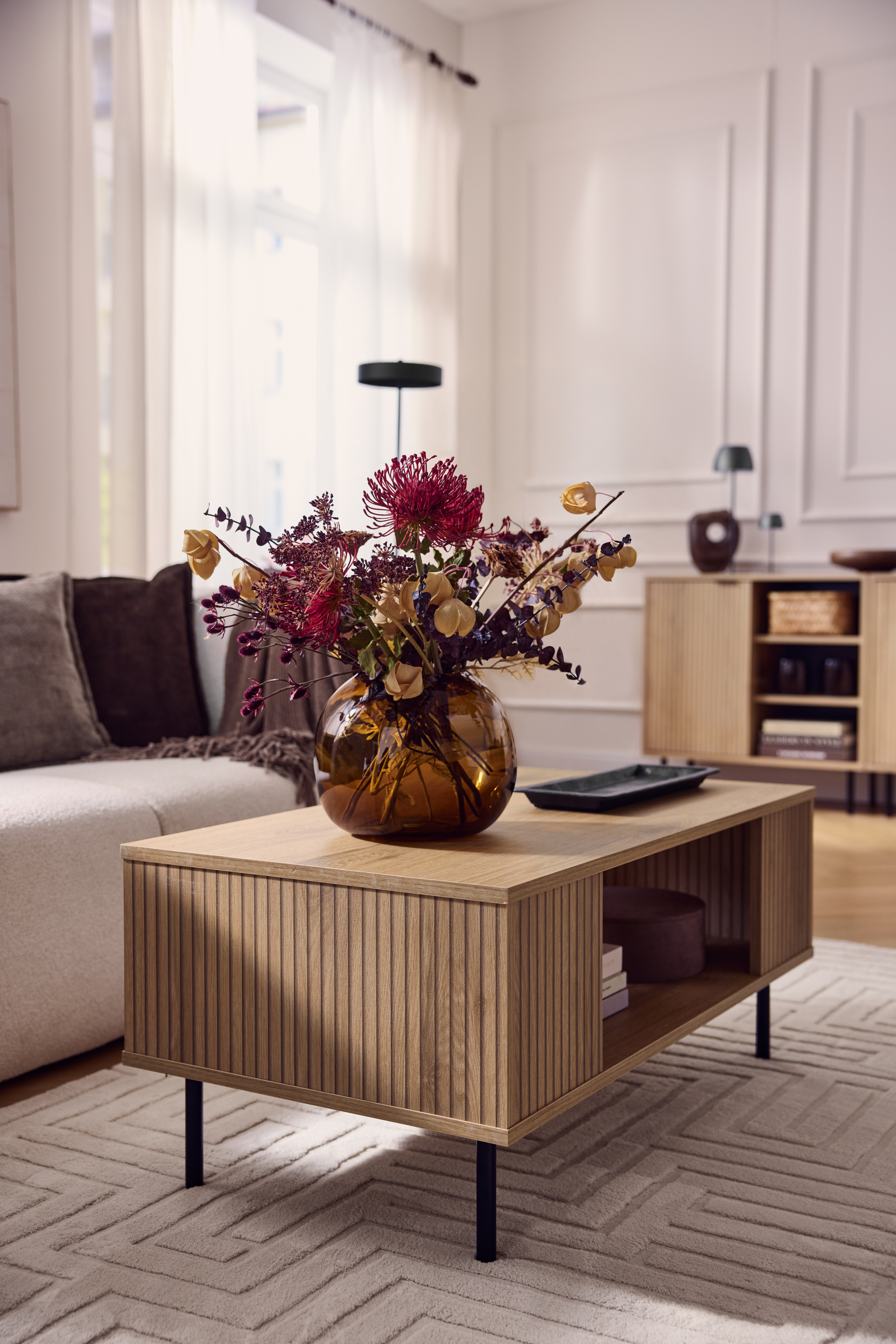 Tavolino da caffè in legno con frontale scanalato, vaso con fiori secchi, divano beige.